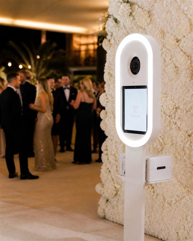 An open-air photo booth kiosk against a floral wall backdrop at a formal event